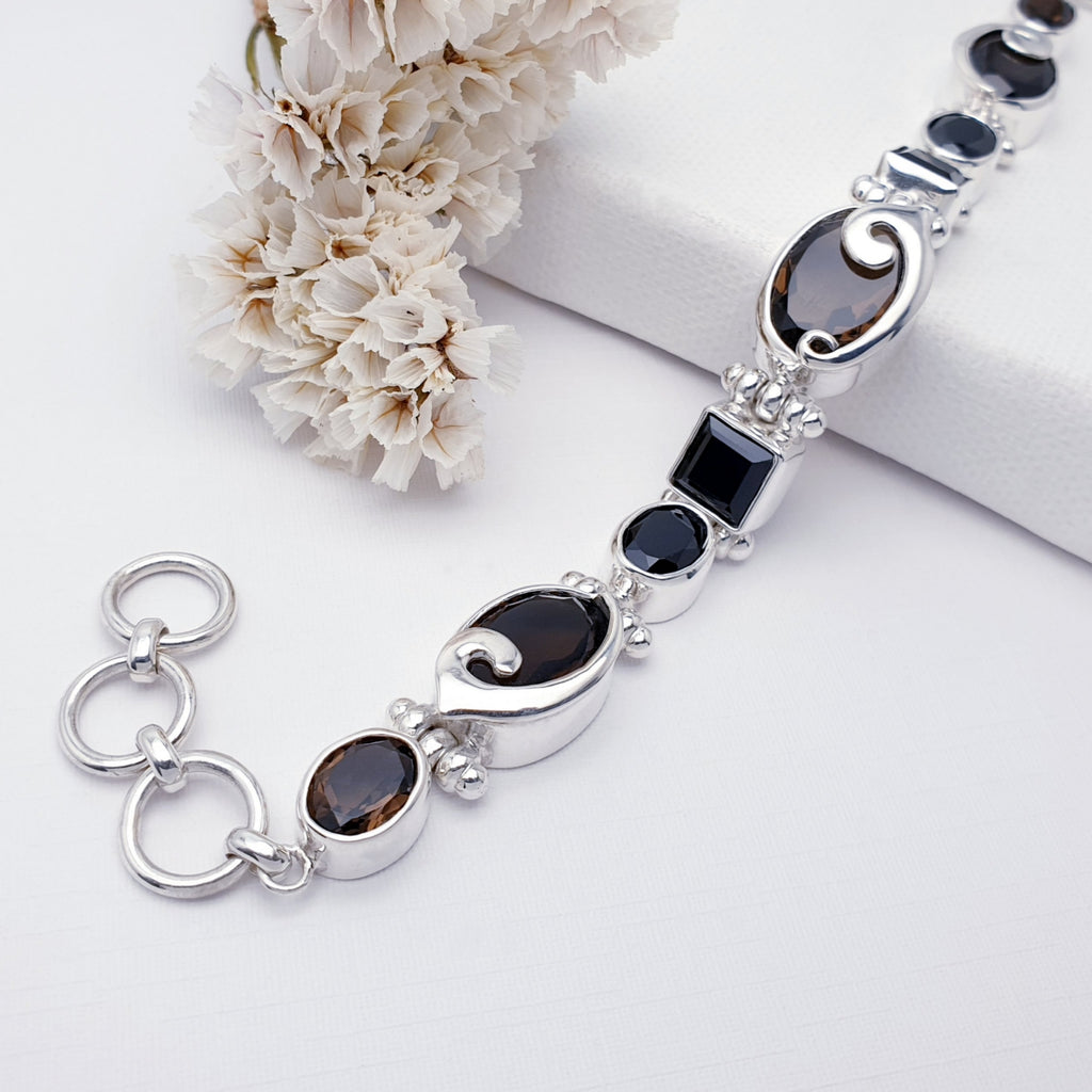 Smoky Quartz and Onyx One-of-a-kind bracelet on a white background with white foliage in the top left corner. The bracelet is also elevated on a white block