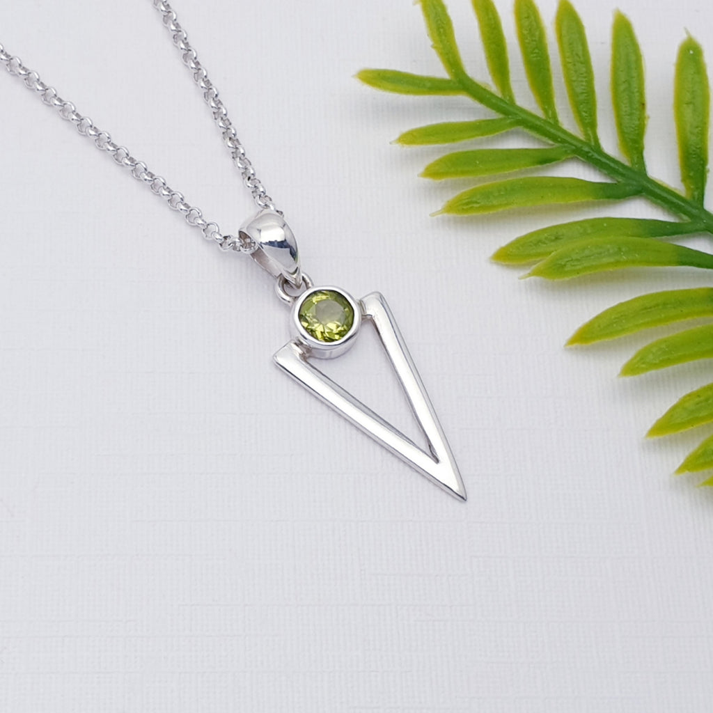 Peridot Acute Triangle Pendant on a white background with green leaves on the right of it.