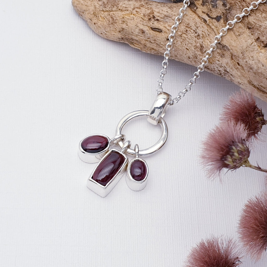 Garnet cluster pendant on a white background, with the chain draped along a piece of drift wood with purple flowers in the bottom right corner