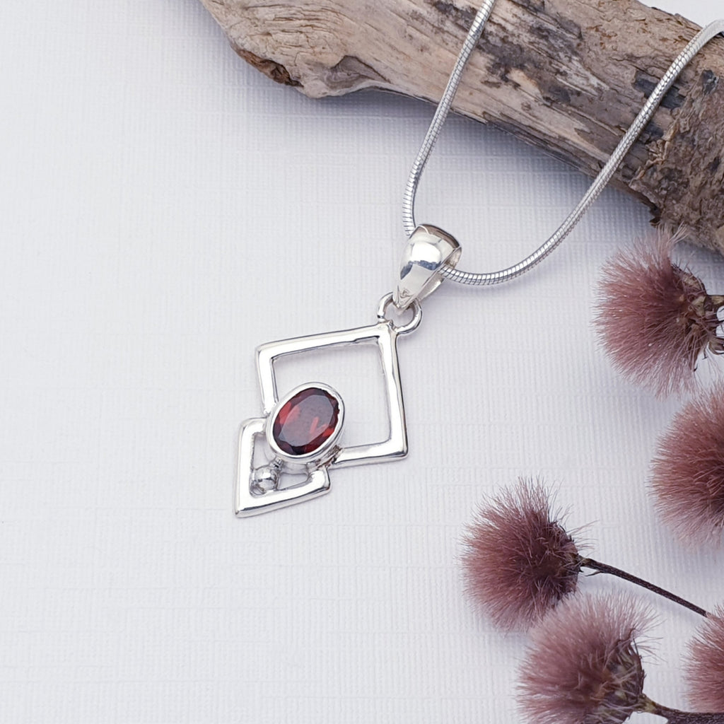 Garnet Triangulum pendant on a white background with burgundy foliage in the bottom right corner. The chain is draped a long a piece of drift wood in the top right corner