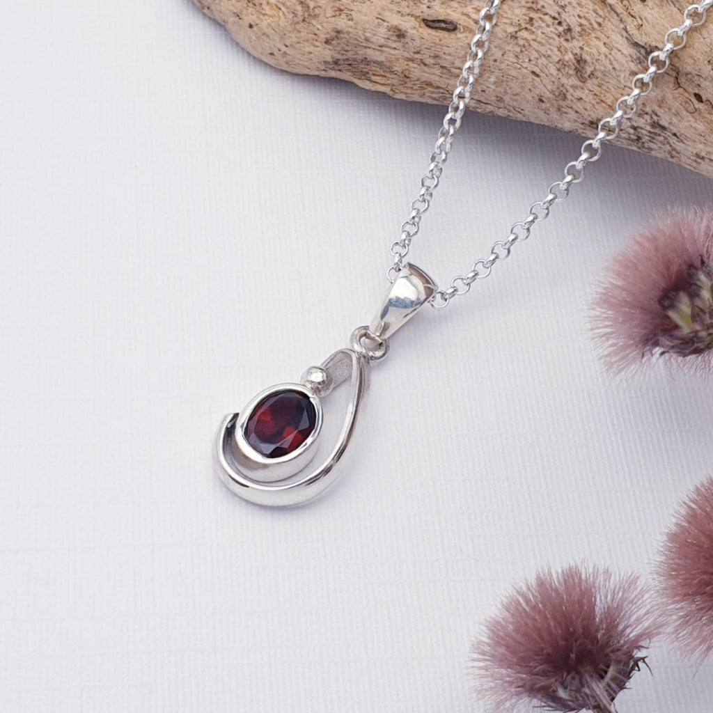 Garnet Midas Pendant on a white background, with a fine belcher chain draped on a piece of drift wood with purple flowers in the bottom right corner