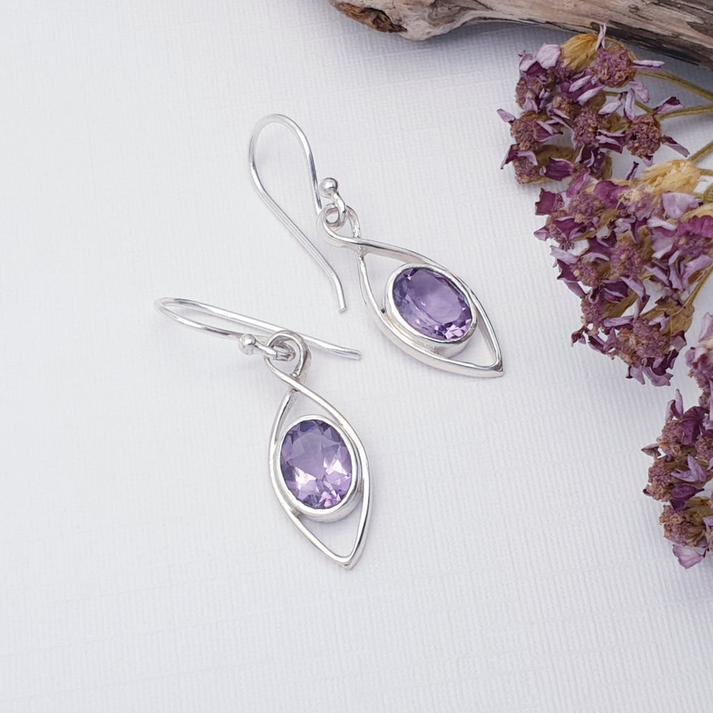 Amethyst Raindrop Earrings on a white background with purple flowers on the right hand side of the image, there is also a piece of drift wood in the top right corner