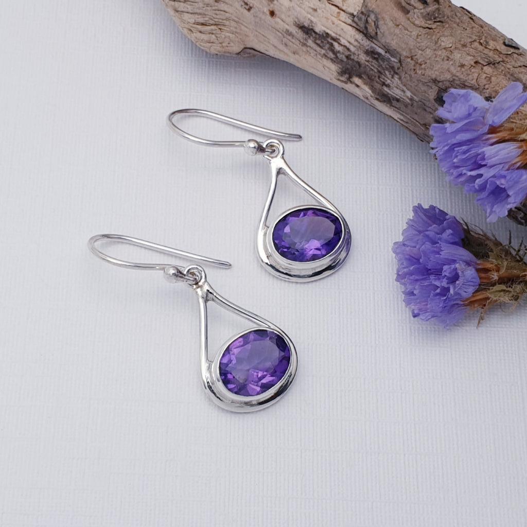 Amethyst Valentina Earrings on a white background with drift wood and purple flowers in the top right hand corner of the image.