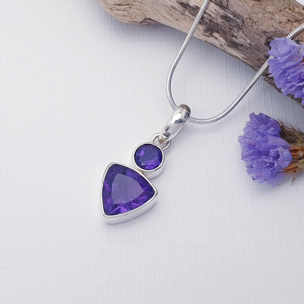 Amethyst Aura pendant on a white background with drift wood and purple flowers in the top right corner of the image