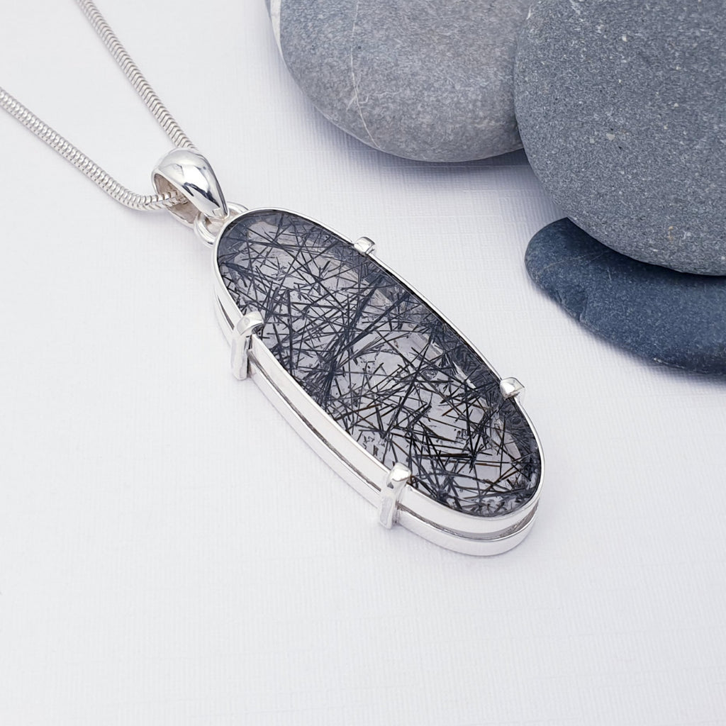 Tourmalinated Quartz Claw Set Oval Pendant on a white background with pebbles in the top right hand corner