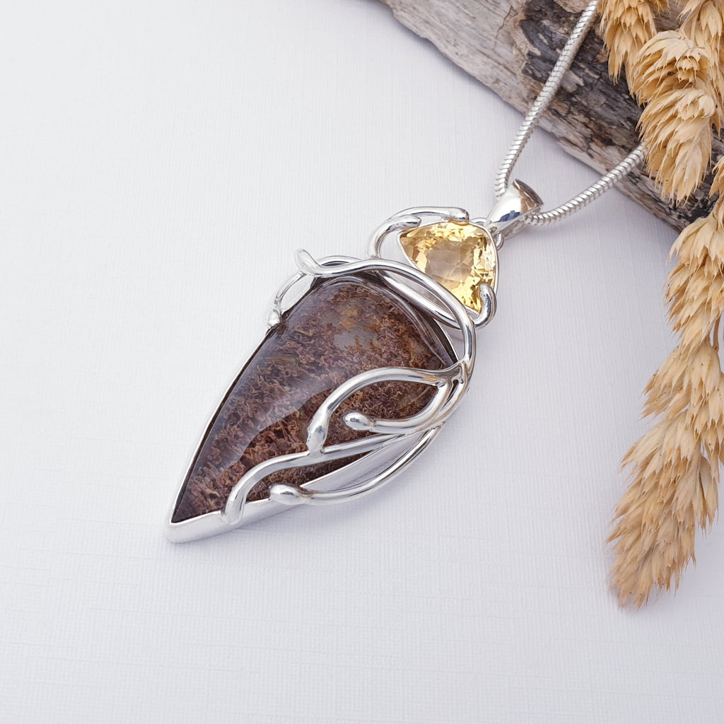 Our Lodolite and Citrine Apollo pendant strung on a heavy snake chain, displayed on a white background with drift wood and barley as decorations