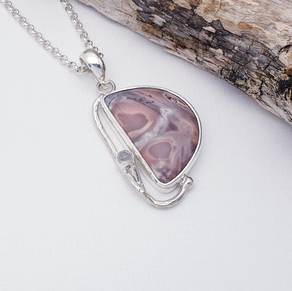 Our Porcelain Jasper and moonstone free form pendant, strung on a medium belcher chain, displayed on a white background with drift wood on the right hand corner as decoration.