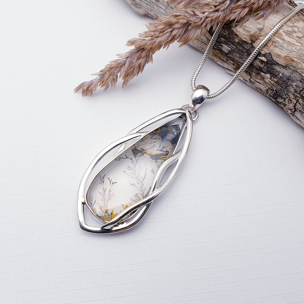 Our One-off Dendritic Quartz Sterling Silver Amalthea Pendant strung on a heavy snake chain, displayed on a white background with drift wood and barley as decorations