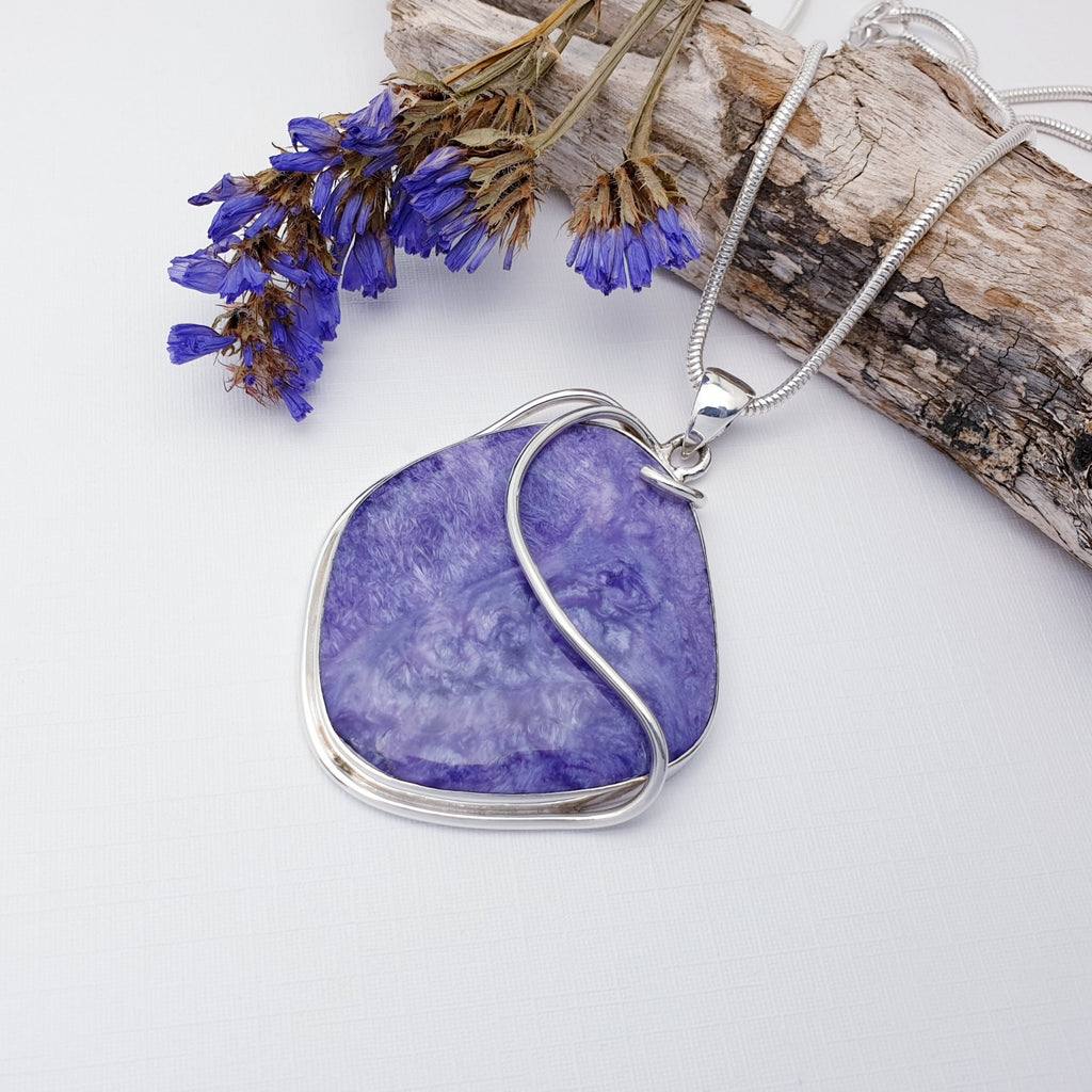 OUR ONE-OFF CHAROITE STERLING SILVER RIBBON PENDANT STRUNG ON A HEAVY SNAKE CHAIN AND DISPLAYED ON A WHITE BACKGROUND WITH PURPLE FLOWERS AND WOOD AS DECORATIONS