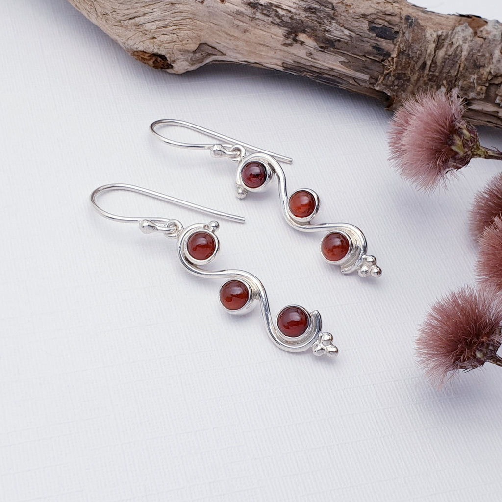 Our garnet sterling silver swirly earrings displayed on a white background with wood and red flowers as decorations