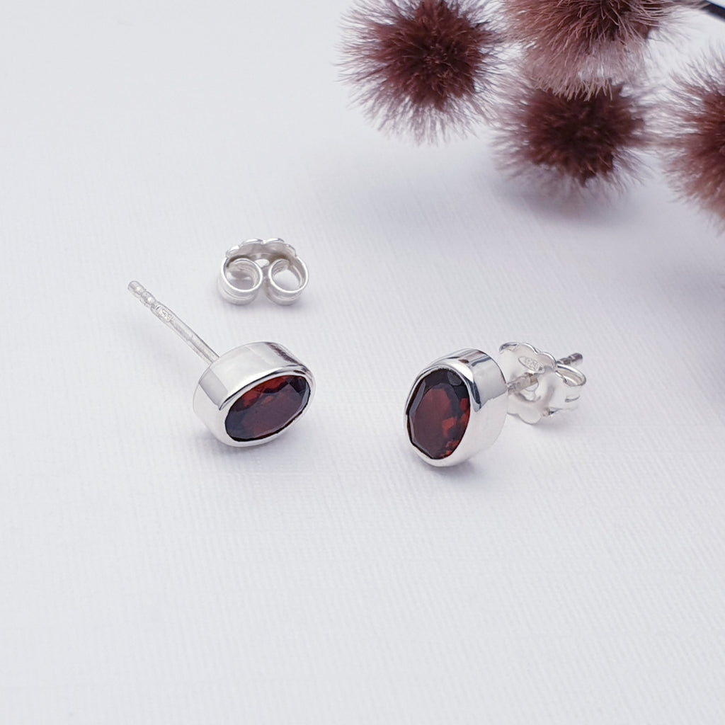 Our simple Garnet Oval studs displayed on a white background with maroon coloured flowers in the background as decoration