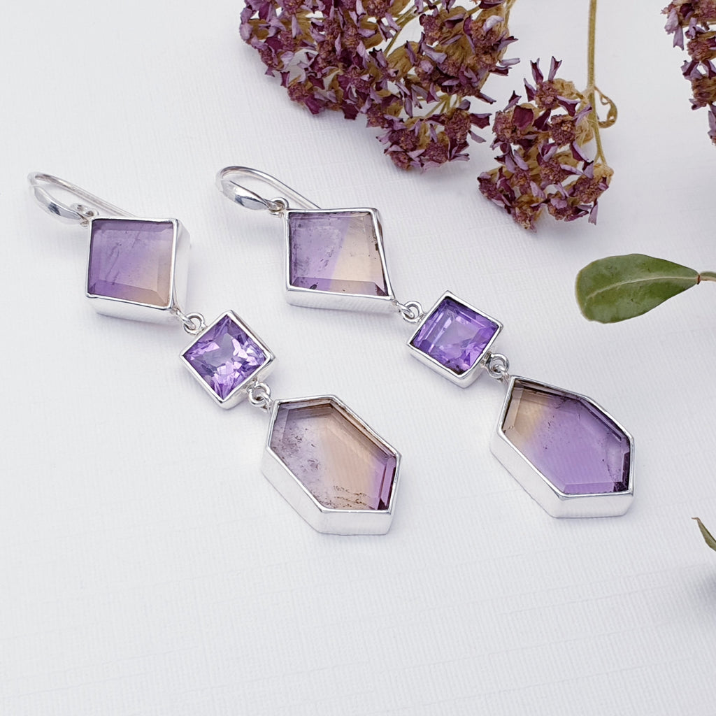 Our one of a kind amethyst and ametrine sterling silver geometric earrings displayed on a white background with purple flowers and a green leaf on the right hand side as decorations