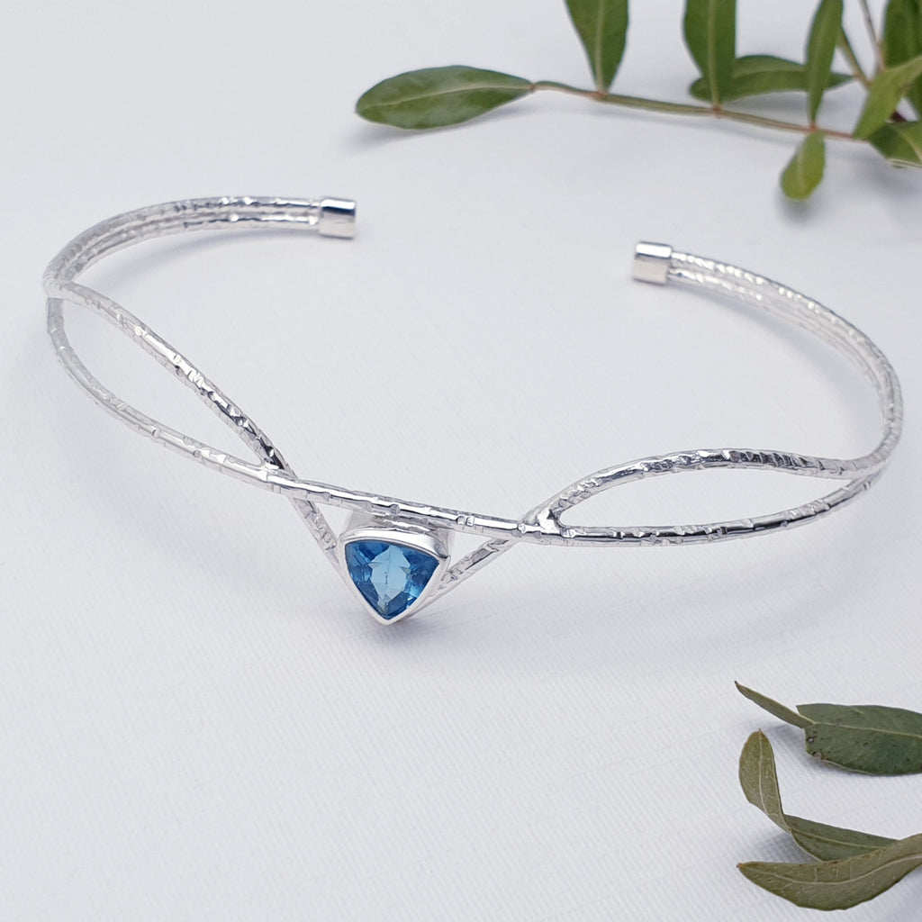 Our blue topaz starling silver galatea cuff displayed on a white background with leafy branches as decoration