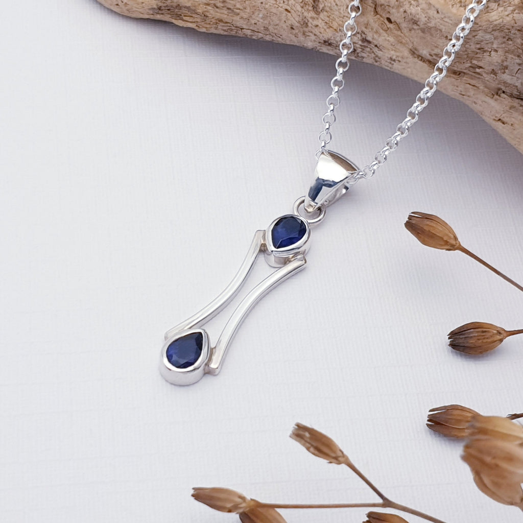 Our Iolite Charis pendant strung on our fine belcher chain, displayed against a white background, with leaves and bark as decorations.