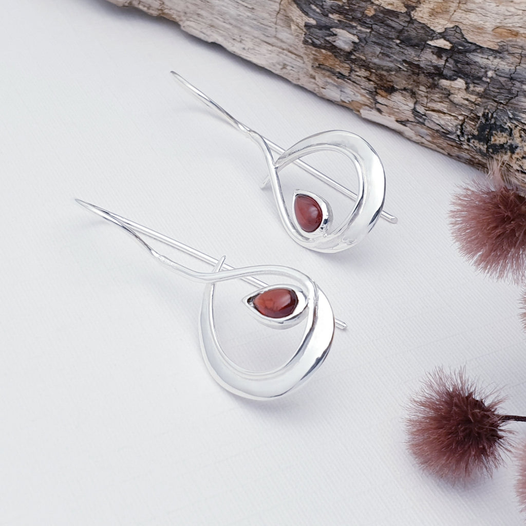 Our Garnet Fluidity earrings displayed against a white background with bark and flowers as decoraton
