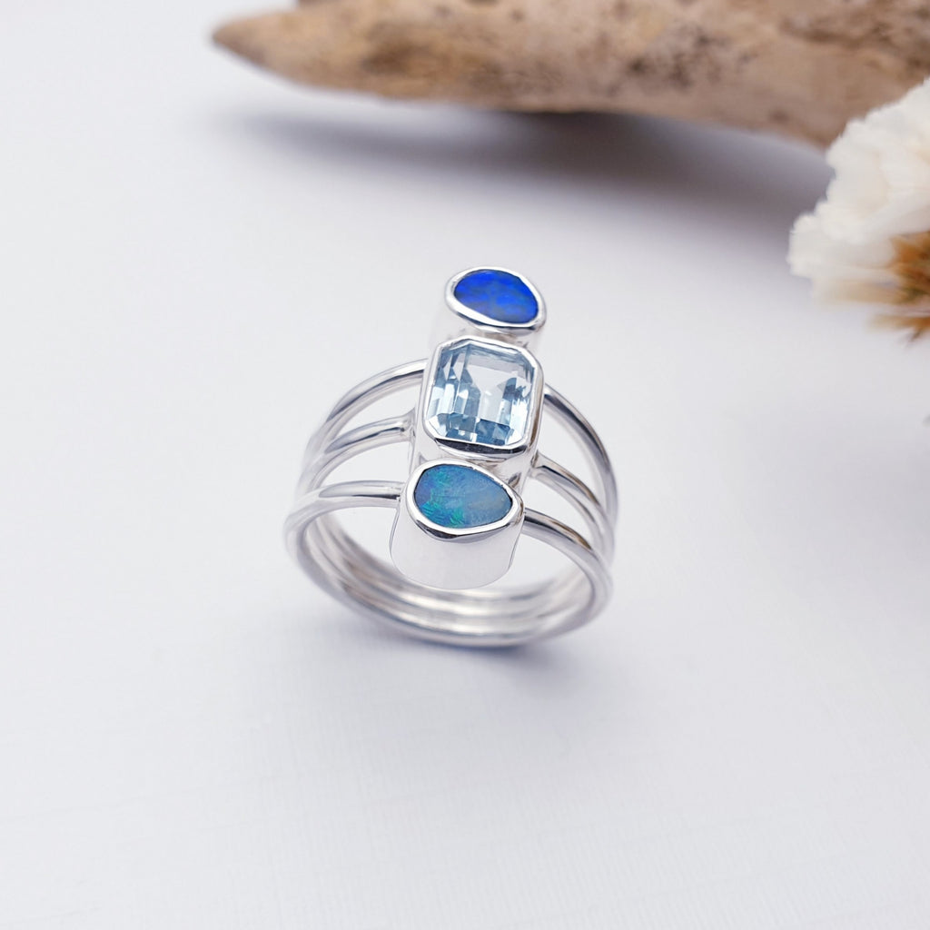Our opal and blue topaz concordia ring against white with driftwood in the background as decoration