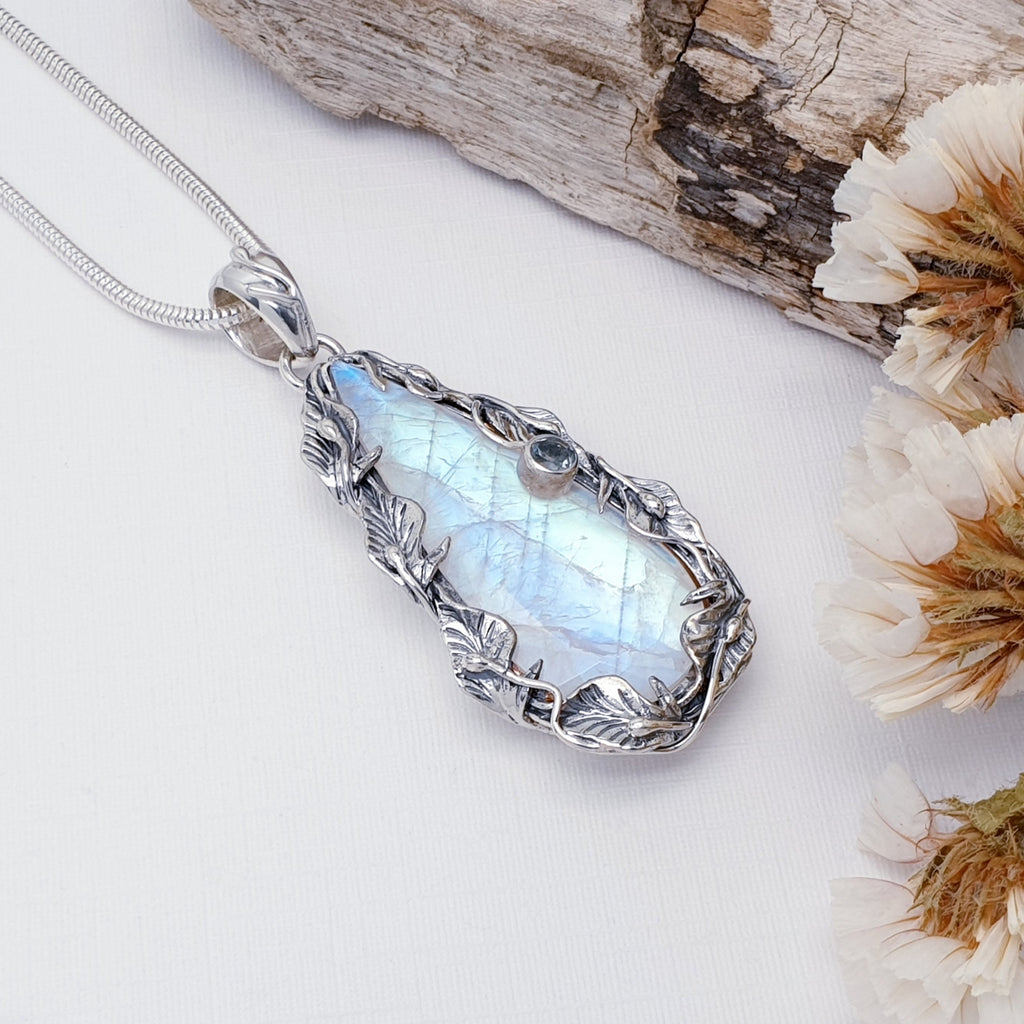 Large silver pendant with Moonstone and Blue Topaz on a white surface with dried flowers and wood.