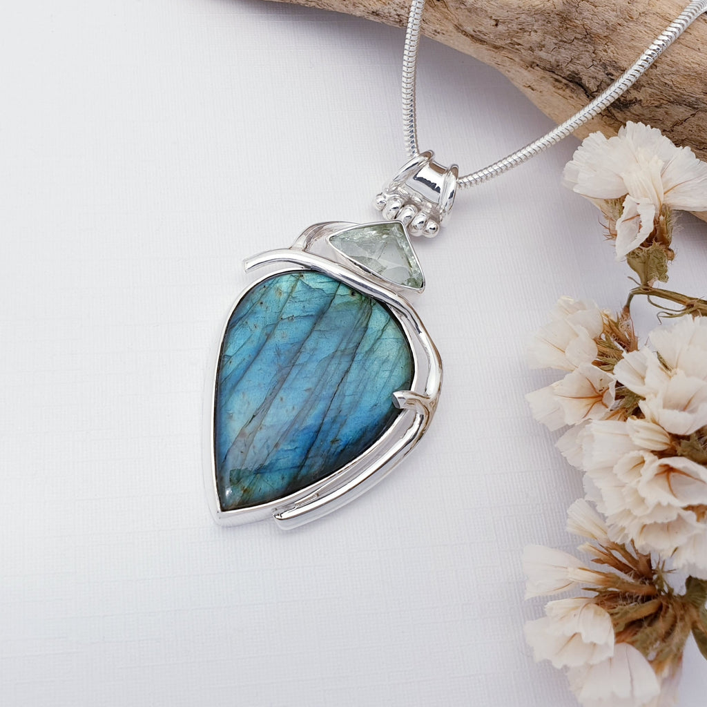 Labradorite and Aquamarine pendant on a white background with wood and flowers on the right hand side
