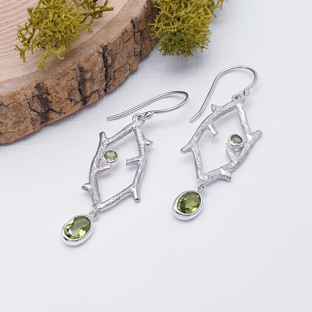 Each earring features an oval, tabletop cut, Peridot stone. The stone elegantly drops down from the bottom of two, interlinked branches, handmade in Sterling Silver. As a finishing touch, a small, round, tabletop cut Peridot stone sits in between the two branches.