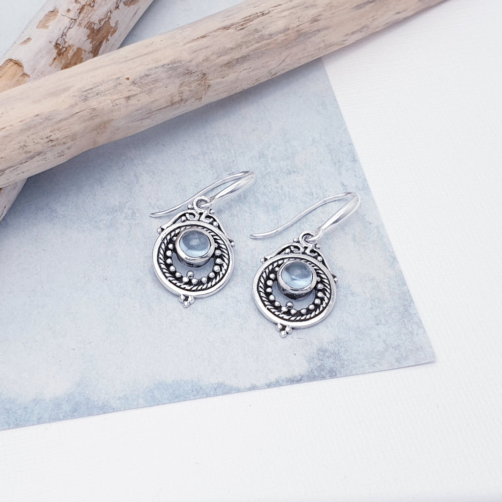 Each earring features a round, cabochon Blue Topaz stone in a simple setting. Intricate Sterling Silver detailing decorates around the stone, and is given depth and presence by the clever use of oxidisation. 