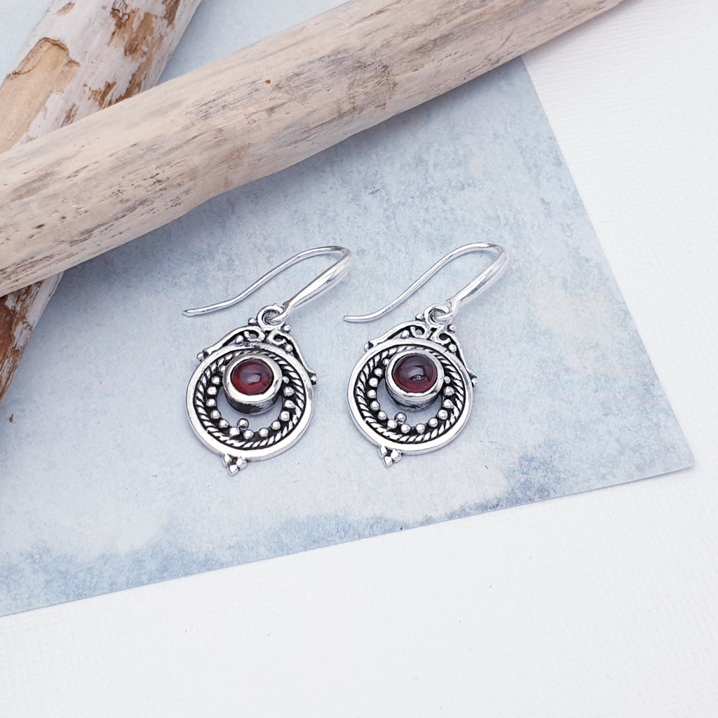 Each earring features a round, cabochon Garnet stone in a simple setting. Intricate Sterling Silver detailing decorates around the stone, and is given depth and presence by the clever use of oxidisation. 
