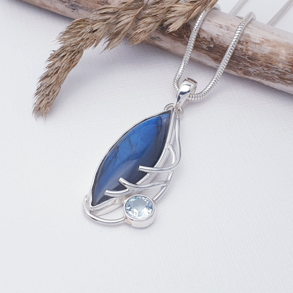 This pendant features a cabochon marquise shaped, Labradorite stone in a simple setting. Inspired by nature, a branch design has been hand worked in Sterling Silver on one side of the stone. A round, tabletop cut Blue Topaz completes the design.