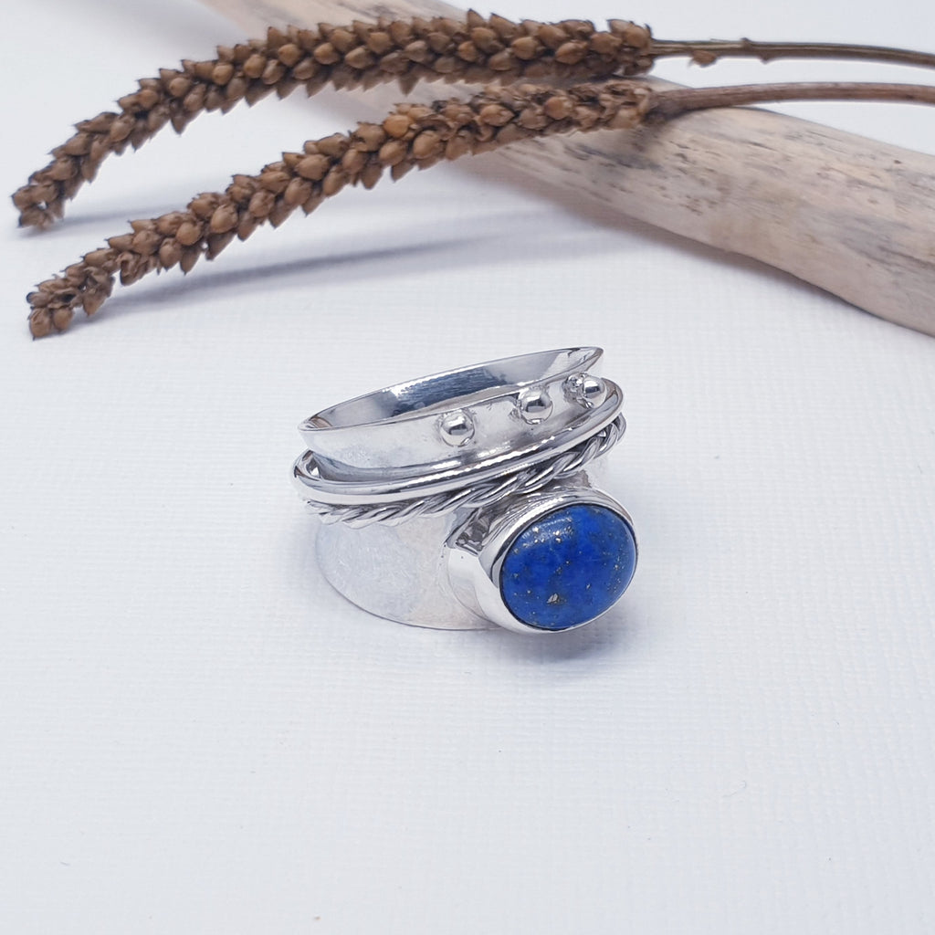 A simple design, this ring features an oval, cabochon, Lapis Lazuli stone set on a wide, slightly concave, Sterling Silver band. Above the stone, two loose thin silver bands, one plain and one twisted, encompass the entirety of the ring. As a finishing touch, above the silver bands our silversmiths have added three small silver balls, adding to the design as well as keeping the silver bands in place. 